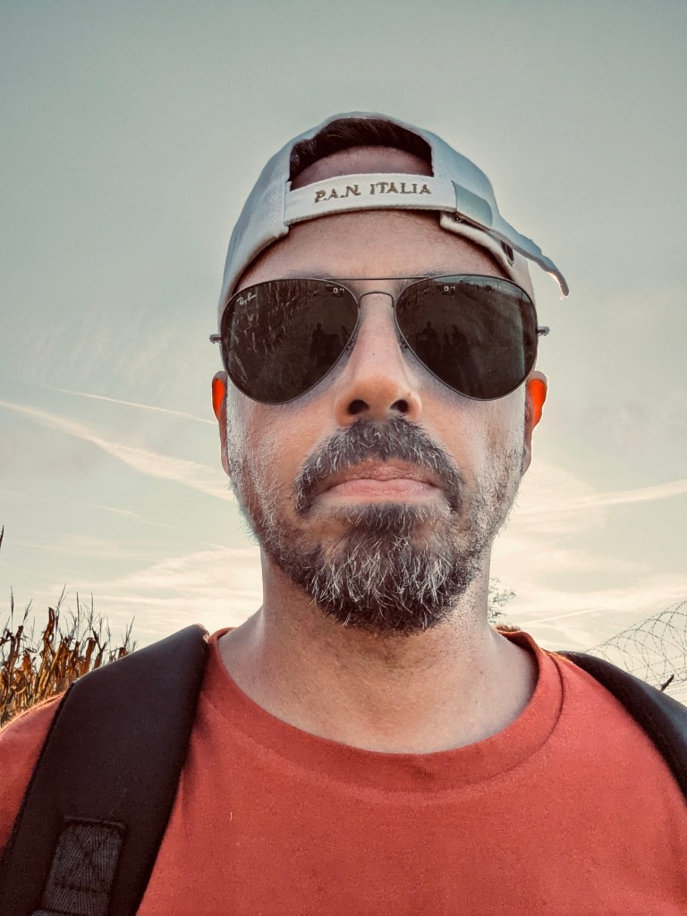 A man wearing sunglasses and a cap, standing outdoors with a field in the background.